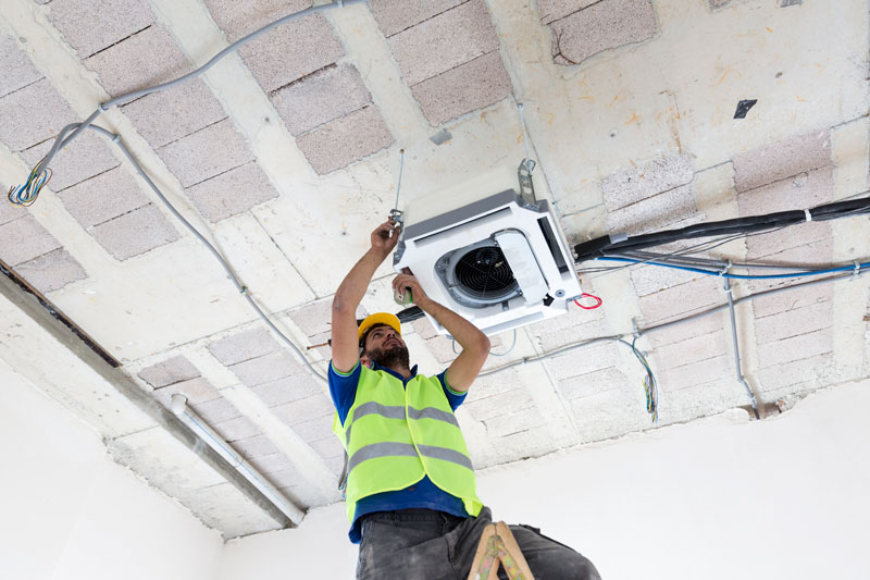 A person wearing a high visibility jacket is performing maintenance to an air conditioning unit.