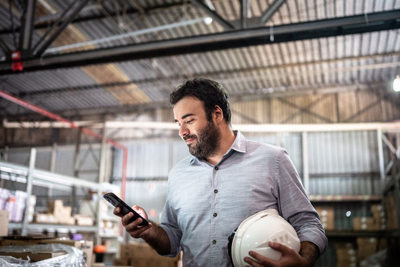 A person is holding protective head gear in one hand and a mobile device in the other, they are looking at the mobile device and smiling.