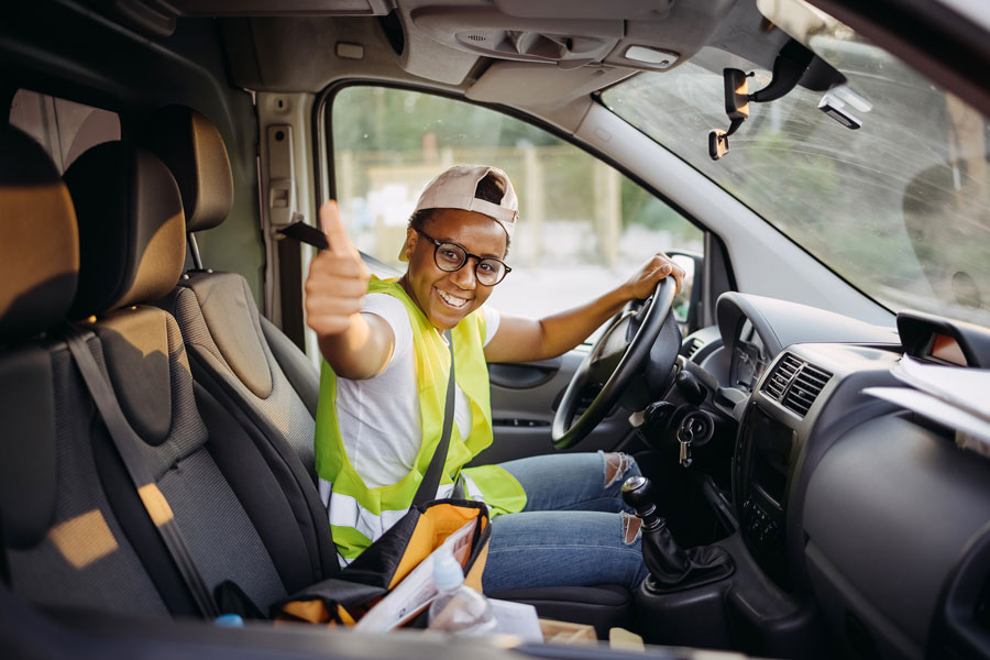 A person wearing a high visibility vest with glasses and a backwards cap, is sitting in a van while smiling and holding one thumb up to the camera.