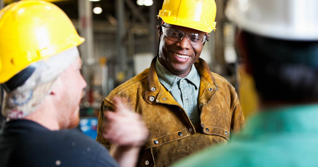 A person is wearing a yellow hard hat and safety goggles and smiling towards another individual wearing a yellow hard hat.