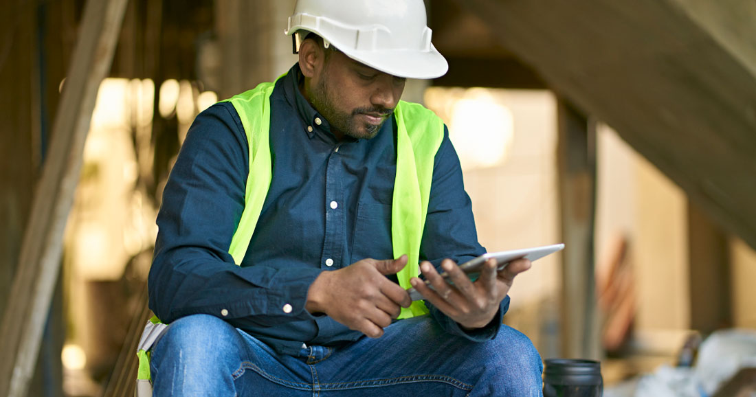 A person is sat down holding a tablet device wearing a high visibility vest and white hard hat