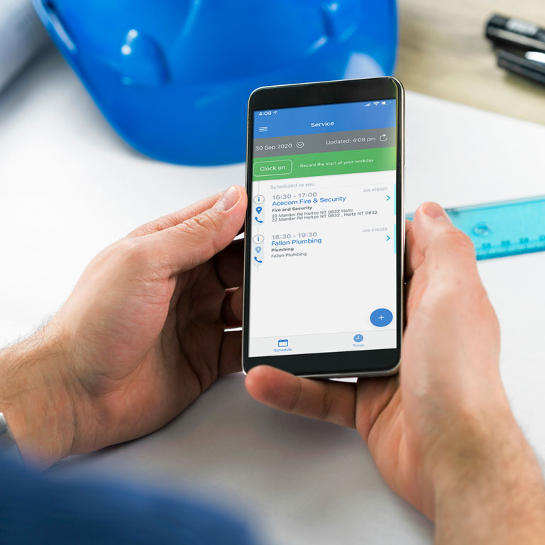 Male hands hold a mobile phone with Simpro Mobile timesheet screen displayed and a blue hardhat and ruler in the background