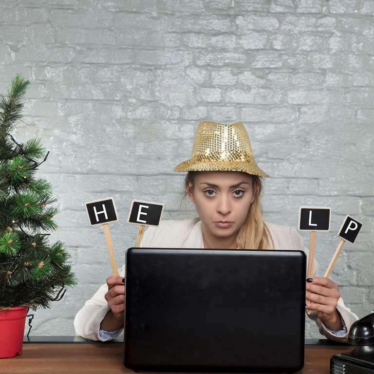 Unhappy woman in an office holds a help sign sits at her laptop surrounded by a small Christmas tree and presents