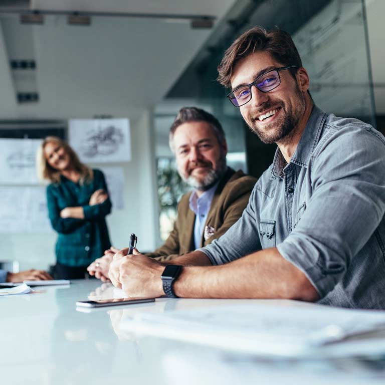 Two businessmen at a table in a modern office smile whilst a female colleague presents financial graphs in the background
