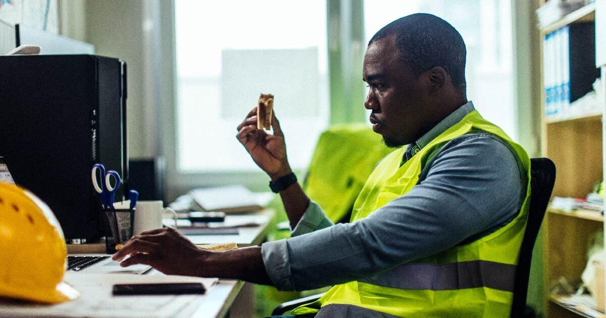 Man in reflective vest eating sandwich and looking at reports on computer
