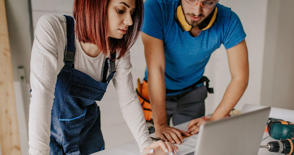 Woman in overalls and man with ear protection looking at computer