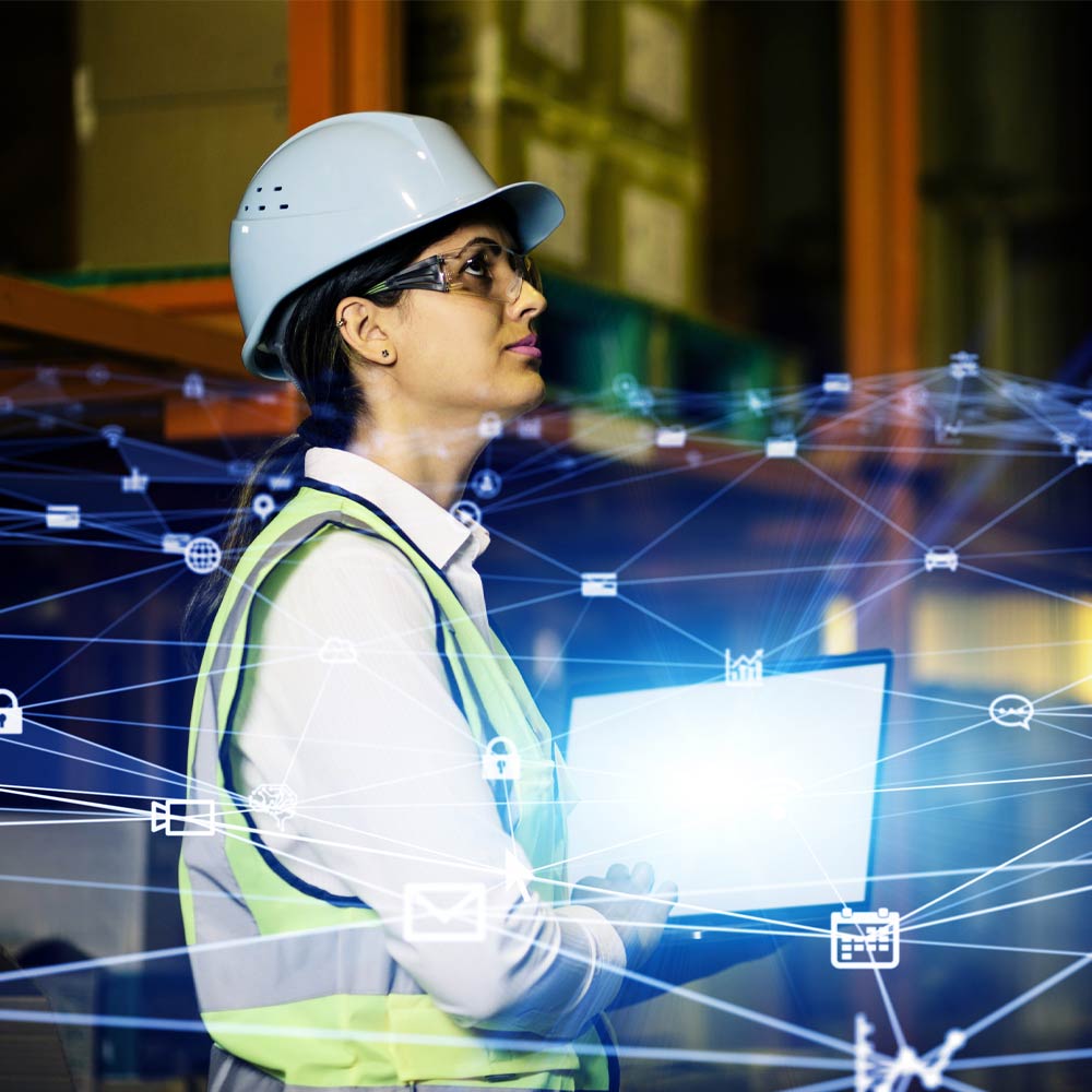 Woman in hard hat and eye goggles holding a laptop looking up in warehouse
