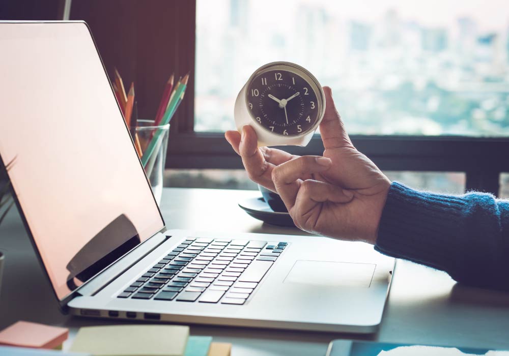 A laptop on a desk and a hand can be seen holding a small clock