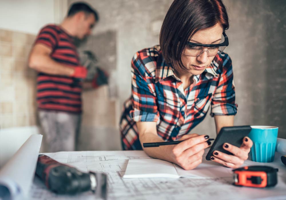 A woman is leaning over a desk holding a tablet device of some sort and in the background a man is using a hammer on a wall