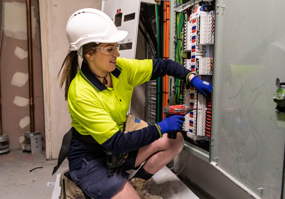 A woman wearing a hardhat is using a hand drill while working on an large electrical device