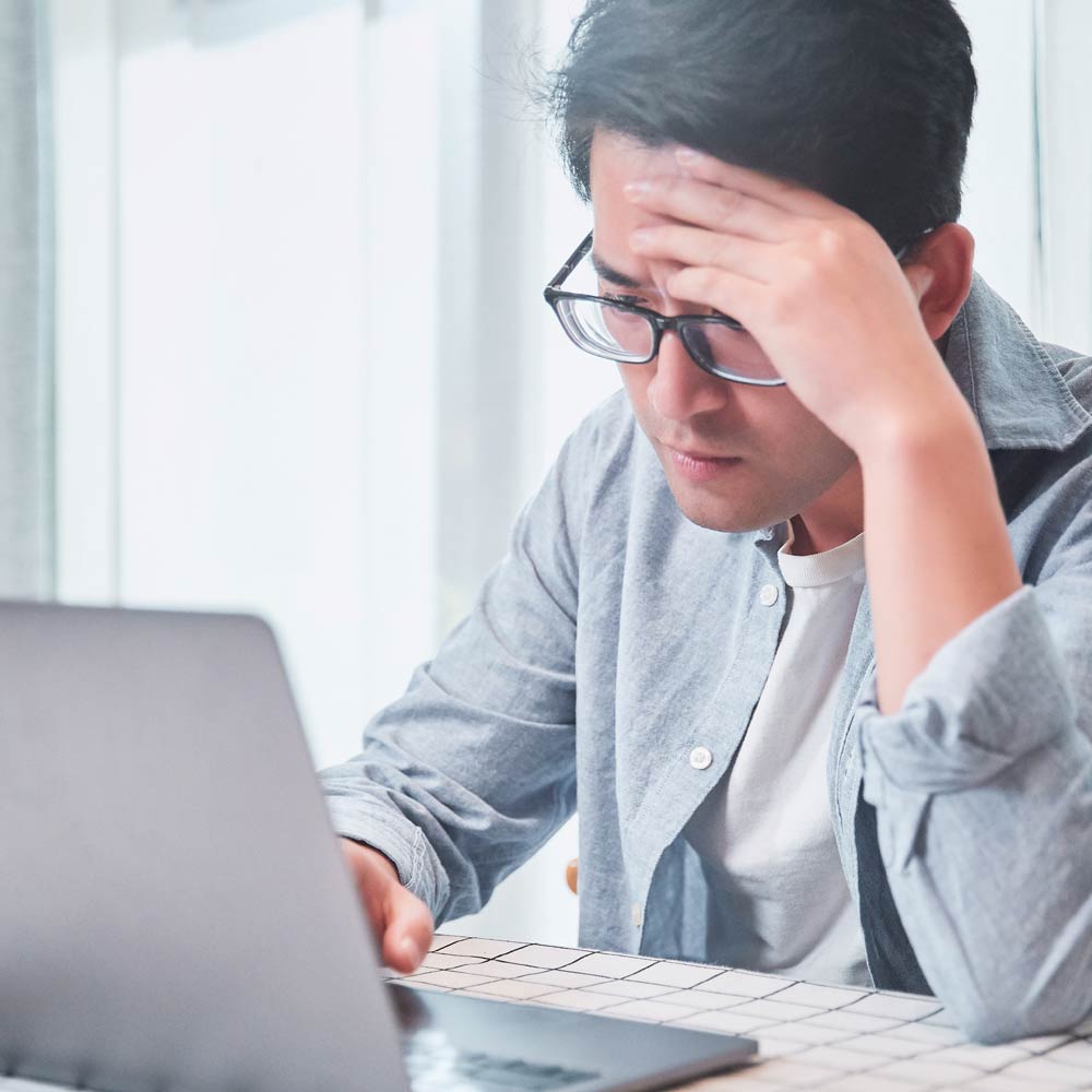 A person is seen sitting at a desk with a laptop with one hand leant on their forhead