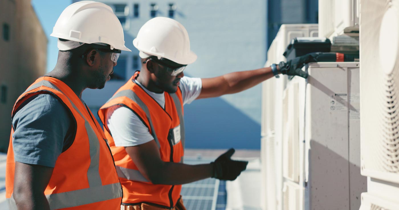 Two men in orange vests and hardhats looking at an HVAC unit