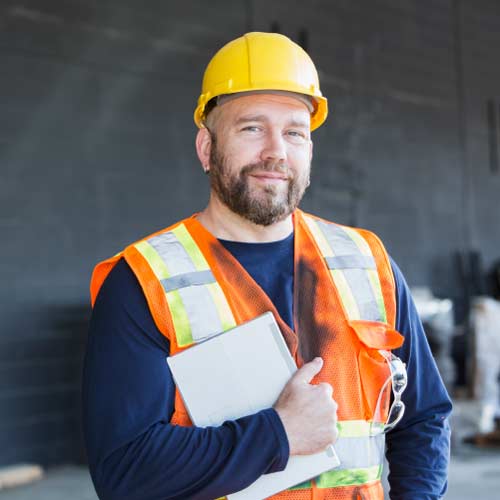 Bearded construction contractor in orange vest wearing yellow hard hat and holding clipboard