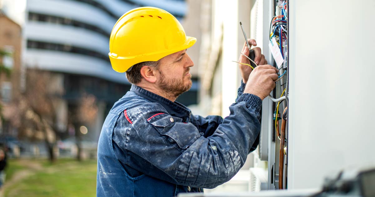 An engineer is wearing a yellow hard hat and blue overalls while working on some electrical wiring