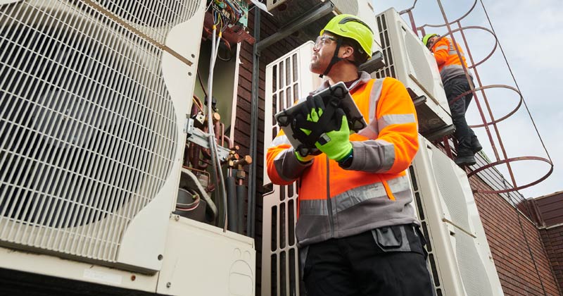 Commercial HVAC installer in yellow hard hat and orange vest looking at HVAC system