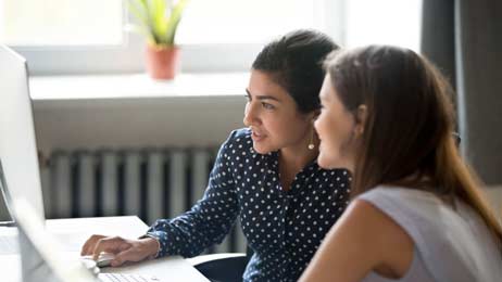 Two professionally dressed women look at a desktop computer while seated at a desk