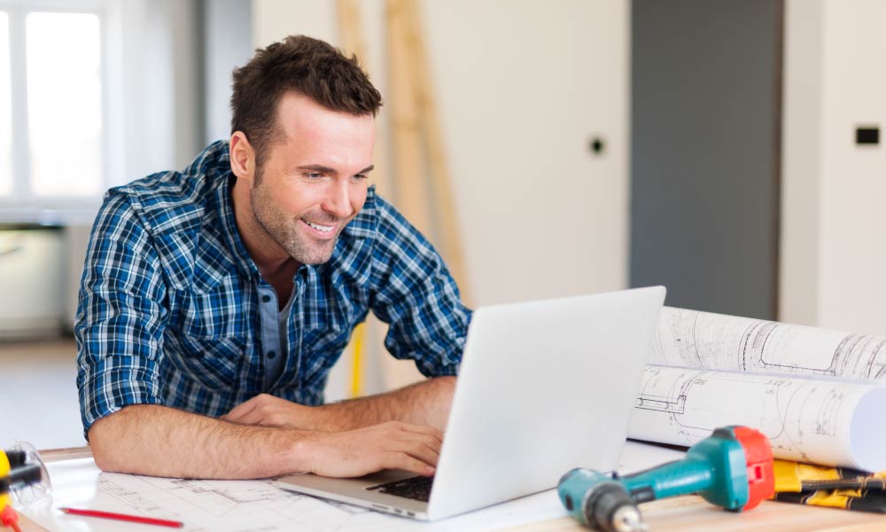 A man is leaning over a desk using a laptop with tools and papers around him