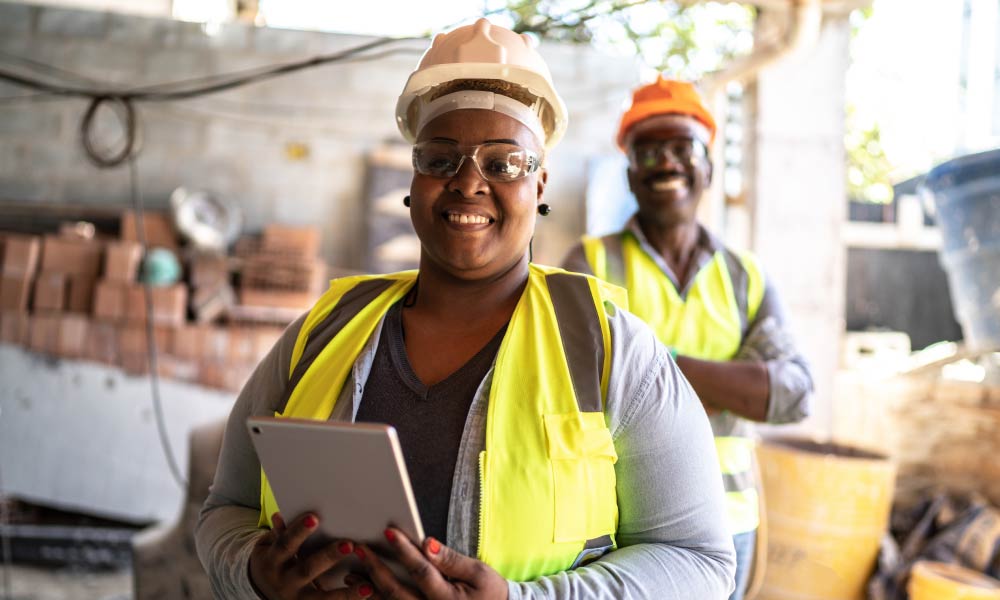 A woman is smiling wearing a high visibility jacket and a hard hat while holding a tablet device