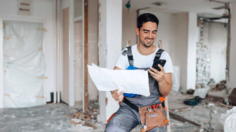 Worker checking phone while holding blueprint in a renovation