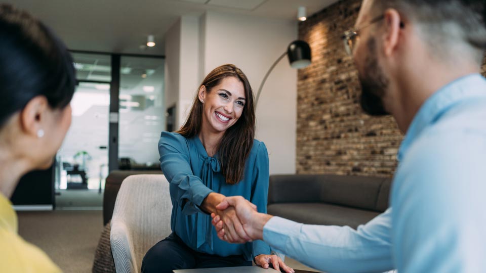 A woman sits in an office space, shaking hands with a man who is sitting across from her beside another woman who is out of frame.