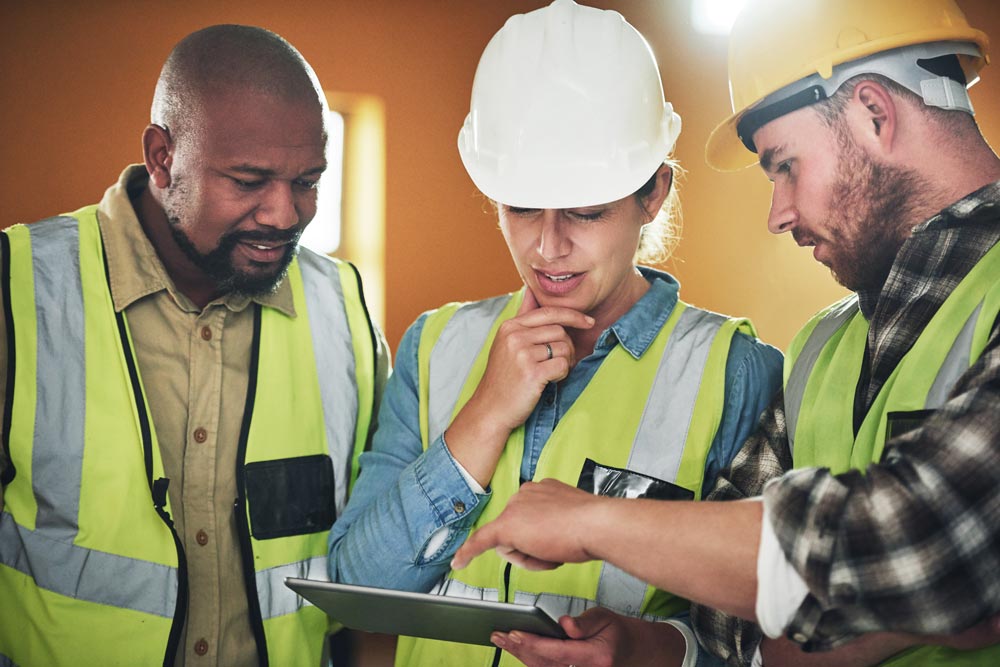 Three people are wearing high visibility jackets and huddled around a tablet device, discussing what they can see on-screen.