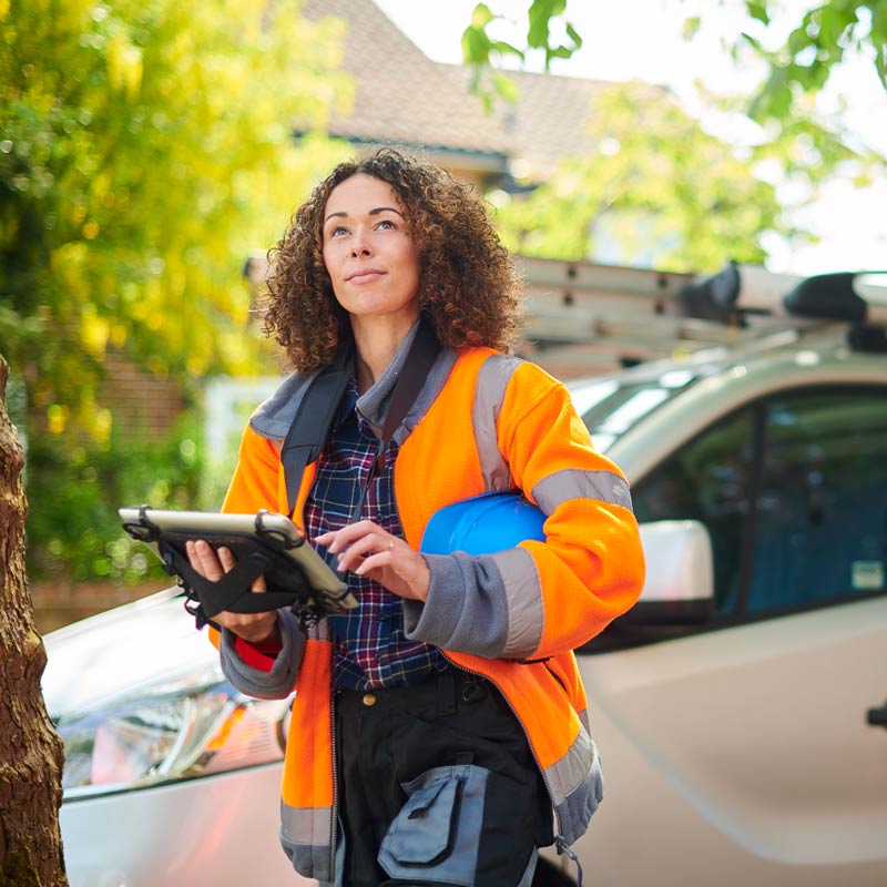 Woman in trade industry wearing orange high visibility jacket and hard hat holding a tablet device with fleet dispatching information and looking upward 