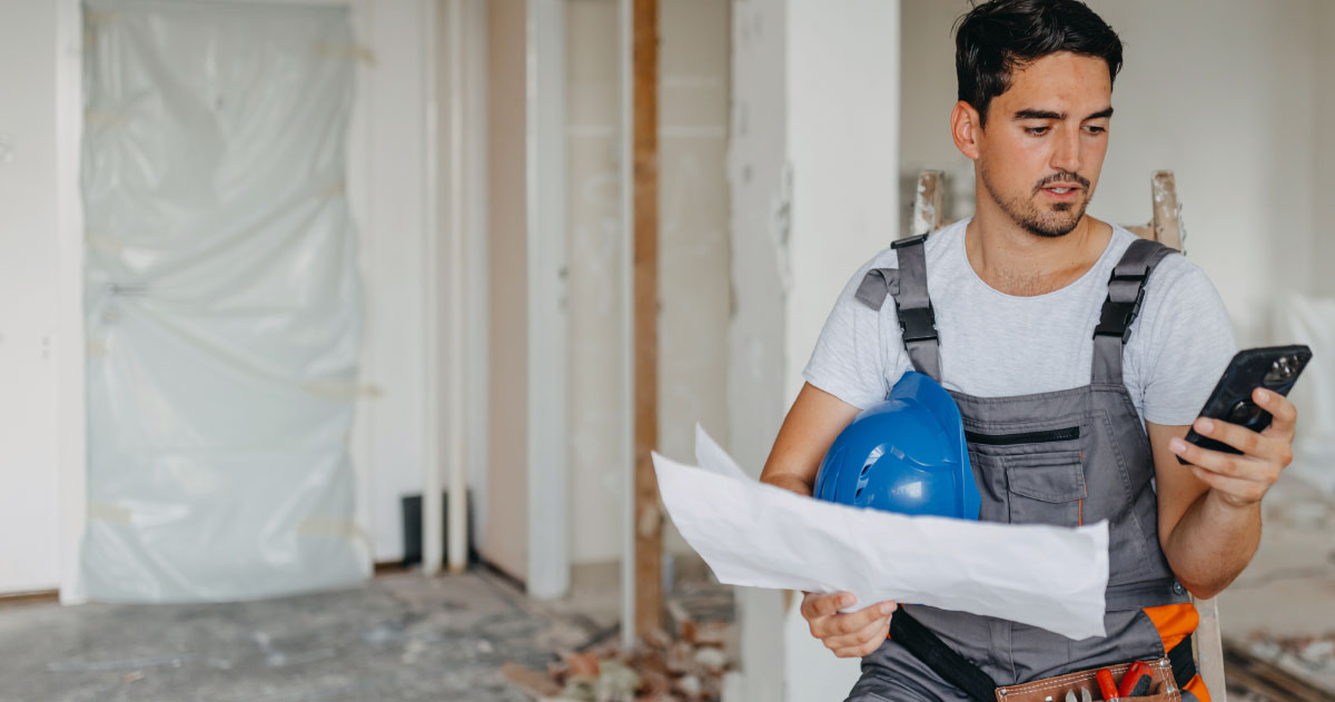 A tradesman is sat on a stool holding a smartphone and a blue hard hat wearing overalls and a toolbelt