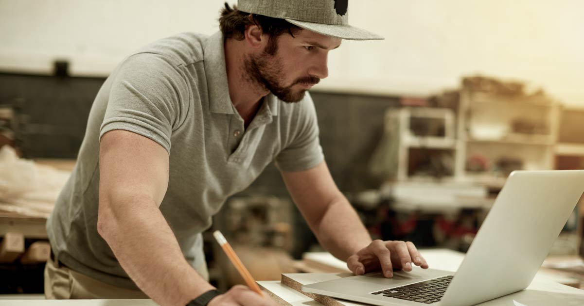 A person wearing a grey polo top and a hat is working at a laptop with a pencil in one hand