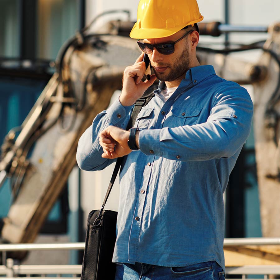 A man is looking at his watch wearing a blue shirt and yellow hard hat