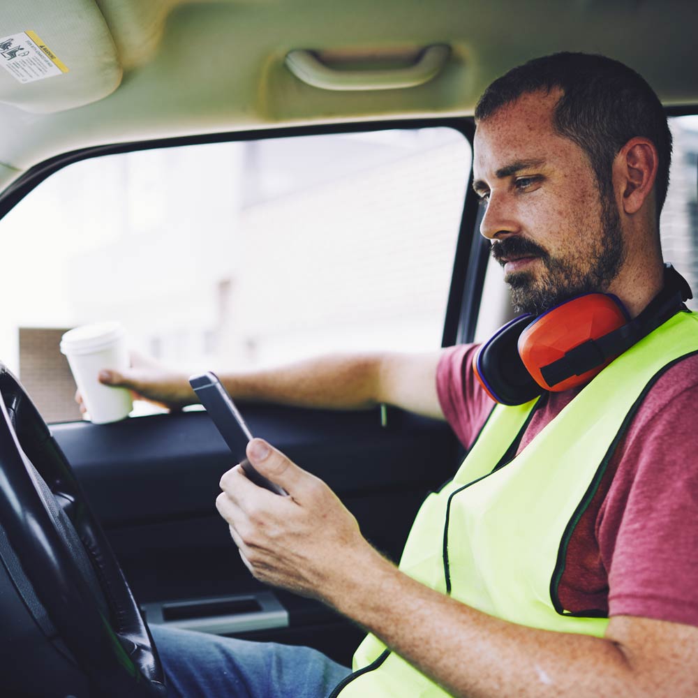 A man is in a car wearing large headphones around his neck and looking at his mobile device
