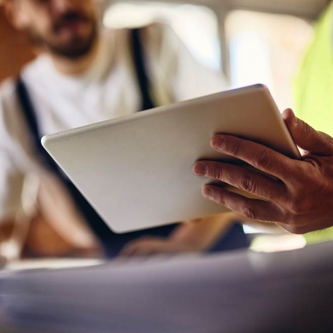 An ipad is held in somebody’s hand with another person out of focus in the background.