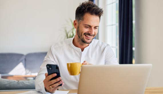 Person hodling mobile device and cup smiling at laptop screen
