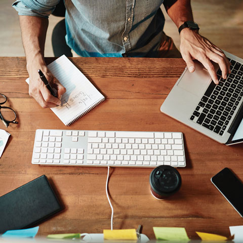 Top down view of user at desk writing notes and operating a laptop